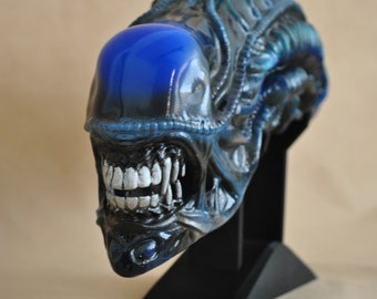 May include: A detailed sculpture of an alien head, featuring a glossy blue dome and intricate, textured details in shades of blue and black. The alien's mouth is open, revealing sharp, white teeth. The sculpture is mounted on a black stand.