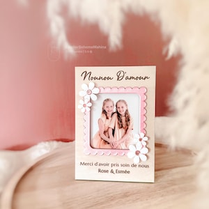 Peut inclure: Un cadre photo en bois avec un bord festonné rose et des décorations de marguerites blanches. Le cadre contient une photo de deux filles et comprend le texte "Nounou D'amour" et "Merci d'avoir pris soin de nous Rose & Esmée."