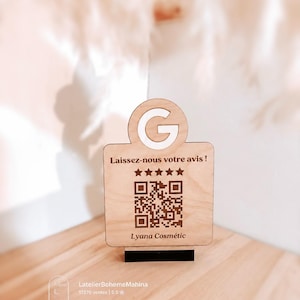 Peut inclure: Un panneau en bois clair avec un sommet circulaire portant la lettre G, le texte Laissez-nous votre avis !, cinq étoiles marron foncé, un QR code carré et le texte Lyana Cosmétic en bas, le tout monté sur un petit socle noir.