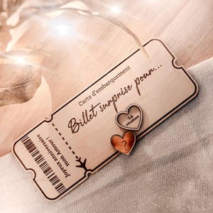 Può includere: Una carta d'imbarco in legno con il testo "Carte d'embarquement Billet surprise pour..." e due piccoli cuori in legno con il testo "La reunion" e un punto interrogativo.