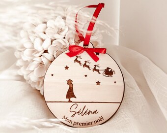 Könnte beinhalten: Ein Weihnachtsbaumschmuck aus Holz mit einem roten Band. Der Schmuck zeigt eine Silhouette eines Mädchens, das zu Santa Claus aufblickt, der in seinem Schlitten mit Rentieren fliegt. Der Text "Selena Mon premier noël" ist in den Schmuck graviert.