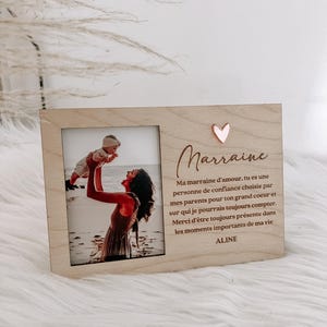Può includere: Una cornice in legno chiaro con una fotografia di una donna che tiene in braccio un bambino. La cornice ha la parola "Marraine" e un ornamento a forma di cuore. È visibile anche del testo sotto la parola "Marraine".