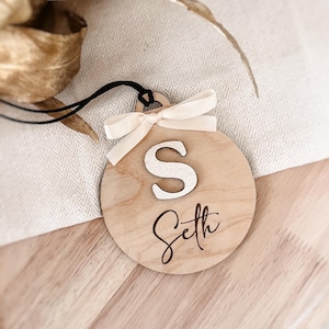 Peut inclure: Une décoration ronde en bois avec une lettre blanche "S" et le nom "Seth" gravé dessus. La décoration est ornée d'un ruban blanc.