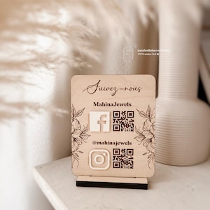 Può includere: Un cartello rettangolare in legno chiaro con base nera che riporta il testo Suivez-nous, MahinaJewels e @mahinajewels. Include icone bianche di Facebook e Instagram, due codici QR e disegni floreali incisi su entrambi i lati.