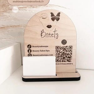 May include: Wooden business card holder with a Beauty Salon Spa design. The holder features a stylized face with a butterfly, the word "Beauty", and social media icons. It includes a QR code and space for cards.