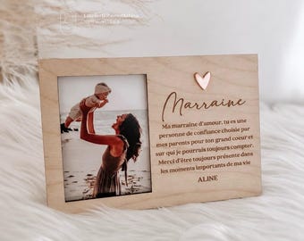 May include: A rectangular light wood photo frame featuring a vertical picture of a person lifting a baby on a beach. The frame includes a small metallic heart icon and French text that reads: Marraine, Ma marraine d'amour, tu es une personne de confiance choisie par mes parents pour ton grand coeur et sur qui je pourrais toujours compter. Merci d'être toujours présente dans les moments importants de ma vie, ALINE.
