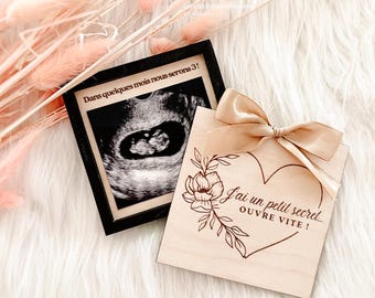 May include: A framed ultrasound image next to a wooden card with a heart design and a bow. The card has the text "I have a little secret, open quickly!" and floral accents. The frame reads "In a few months we will be 3!"