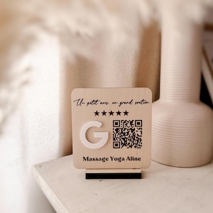 Può includere: Un cartello quadrato in legno con base nera che presenta il testo "Un petit avis, un grand soutien," cinque stelle nere, una lettera "G" bianca, un codice QR nero e il testo "Massage Yoga Aline" stampato in nero sulla superficie in legno chiaro.