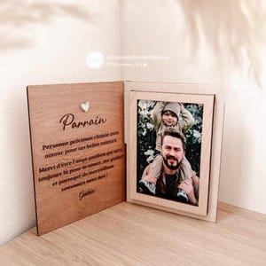 Può includere: Una cornice in legno aperta, che mostra una foto di un uomo e un bambino. Presenta una targa in legno con la parola "Parrain", un messaggio in francese e il nome "Gabin".