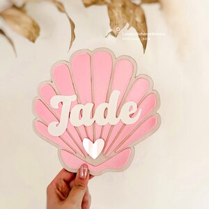 Peut inclure: Décoration en forme de coquillage rose avec le nom "Jade" en écriture blanche. Un petit cœur blanc est au centre. Le coquillage est tenu par une main.