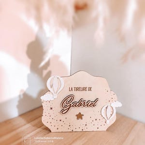 Peut inclure: Une tirelire en bois clair aux bords festonnés, portant l'inscription 'LA TIRELIRE DE Gabriel' en lettres sombres. Le motif comprend deux montgolfières blanches, de petits nuages, un motif de minuscules étoiles et une découpe en forme d'étoile.