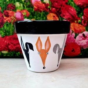 May include: A white ceramic flower pot with a black rim. The pot is decorated with three painted animal faces: a black dog, an orange fox, and a grey dog. The faces are all looking out from the pot.
