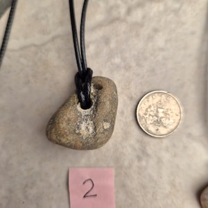 May include: A natural stone pendant necklace with a black cord. The stone is a light brown color with a hole drilled through it. A silver coin and a pink sticky note with the number 2 are also in the image.
