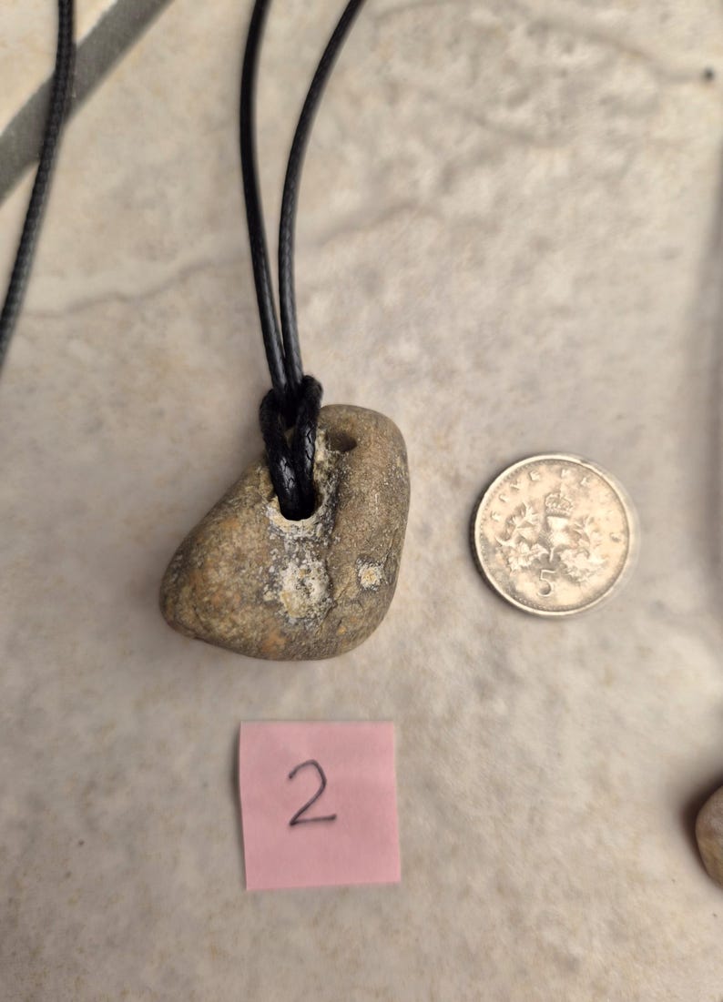 May include: A natural stone pendant necklace with a black cord. The stone is a light brown color with a hole drilled through it. A silver coin and a pink sticky note with the number 2 are also in the image.