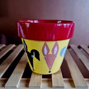 May include: A yellow terracotta flower pot with a red rim. The pot is painted with three animal faces: a black dog, a pink rabbit, and a blue cat.