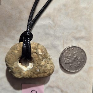 May include: A natural stone pendant with a hole, strung on a black cord. A five-cent coin is next to the pendant for size comparison. The stone is beige with a lighter interior. A pink tag with the number 8 is also visible.