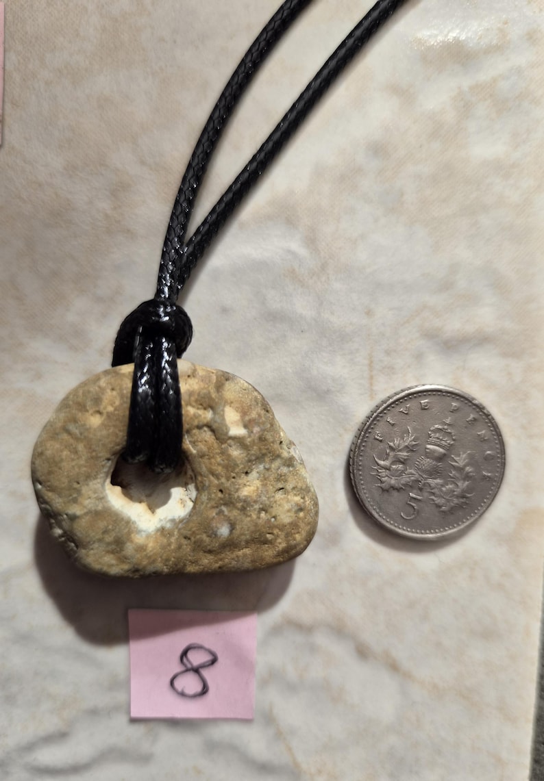 May include: A natural stone pendant with a hole, strung on a black cord. A five-cent coin is next to the pendant for size comparison. The stone is beige with a lighter interior. A pink tag with the number 8 is also visible.