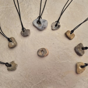 May include: A collection of stone pendants on black cords. The pendants vary in shape and color, ranging from light tan to gray. Each stone has a hole for the cord to pass through, creating a natural, earthy aesthetic. The pendants are arranged on a neutral surface.