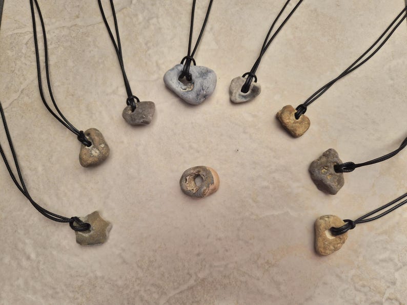 May include: A collection of stone pendants on black cords. The pendants vary in shape and color, ranging from light tan to gray. Each stone has a hole for the cord to pass through, creating a natural, earthy aesthetic. The pendants are arranged on a neutral surface.