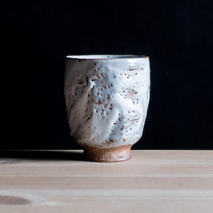May include: A ceramic cup with a textured, off-white glaze featuring small brown speckles and indentations. The vessel has a cylindrical shape with a slightly tapered base that reveals the raw, unglazed brown clay material at the bottom.