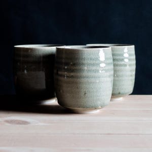 May include: Three cylindrical ceramic cups featuring a light sage green glaze with a visible crackle finish. Each cup has a slightly ribbed texture along the body and a smooth, rounded rim, standing on a small, plain white circular base.