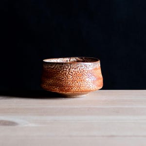 Può includere: Una piccola ciotola in ceramica rotonda con una superficie ruvida e screpolata nei toni dell'arancione, marrone e beige. La ciotola presenta un bordo ampio e leggermente irregolare e una base affusolata, con una finitura rustica in argilla.