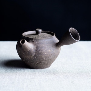 Puede incluir: Tetera de cerámica marrón oscuro con tapa y boquilla. La tetera tiene un cuerpo redondeado y una superficie texturizada. La boquilla está en ángulo y tiene una abertura ancha. La tapa tiene un pequeño pomo en la parte superior.