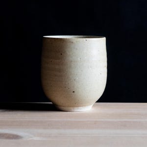 Könnte beinhalten: Ein zylindrischer Keramikbecher mit gesprenkelter beiger Oberfläche und einem glatten, abgerundeten Körper. Das Gefäß verfügt über einen dezent verjüngten Boden und einen schlichten Rand, was eine natürliche, erdige Textur auf der Außenseite zeigt.