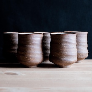 May include: A set of five brown ceramic cups with a textured, wavy surface and a matte finish. Each cup features a cylindrical shape that tapers slightly towards the base, showcasing a rustic, earthy appearance with visible clay grain and subtle variations.