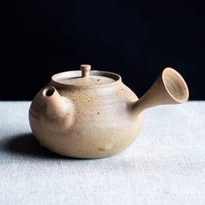 Può includere: Una teiera in ceramica beige con un corpo arrotondato, un beccuccio e un coperchio con una piccola maniglia. La teiera ha una consistenza macchiettata e una finitura opaca. Il beccuccio è angolato verso l'alto. Lo sfondo è nero.
