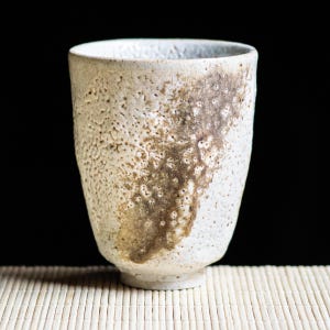 Può includere: Una tazza in ceramica fatta a mano con un esterno bianco sporco strutturato e un design marrone screziato. La tazza ha un bordo leggermente svasato e una base arrotondata. La tazza è alta circa 10 cm.