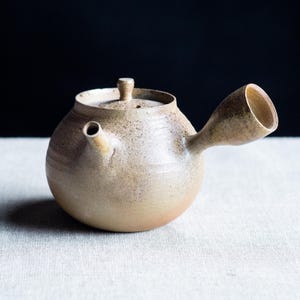 Puede incluir: Una tetera de cerámica hecha a mano, con un cuerpo redondeado y un pico corto e inclinado. La tetera es de color beige moteado con una tapa y un asa pequeña. El fondo es oscuro y la tetera está sobre una superficie blanca.