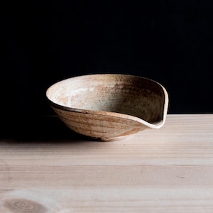 Może przedstawiać: Ceramiczna miska o nakrapianym beżowo-brązowym wykończeniu, posiadająca szeroką, płytką czaszę oraz wyraźny dzióbek do nalewania z jednej strony. Miska ma teksturowany, ziemisty wygląd z subtelnymi różnicami w odcieniach na całej powierzchni.