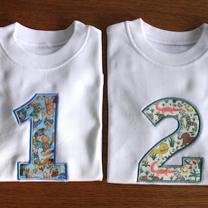 May include: Two white short-sleeved t-shirts featuring large appliquéd numbers on the front. The left shirt displays the number one with a blue border and a colourful animal print pattern. The right shirt shows the number two with a green border.