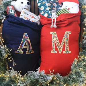 May include: A navy blue and a red fabric sack, each featuring a large embroidered letter on the front. The navy sack displays the letter A, and the red sack displays the letter M. Both letters are filled with a colourful holiday-themed pattern.