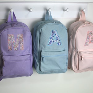 May include: Three backpacks in lavender, light blue, and pale pink, each featuring a front zipped pocket and a large fabric letter appliqué on the main compartment. The letters M, A, and A are decorated with a colourful floral pattern.
