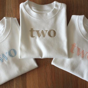 May include: Three white t-shirts with the word "two" embroidered on the front in different colours: light blue, gold, and pink.