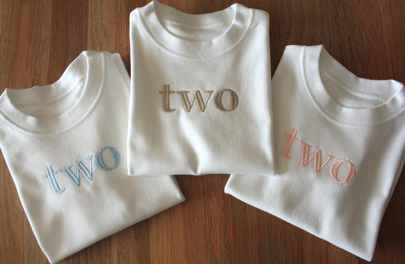 May include: Three white t-shirts with the word "two" embroidered on the front in different colours: light blue, gold, and pink.