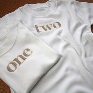 May include: Two white t-shirts with the words "one" and "two" embroidered in gold on the front.