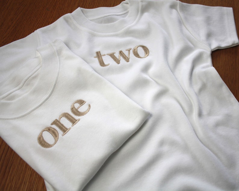 May include: Two white t-shirts with the words "one" and "two" embroidered in gold on the front.