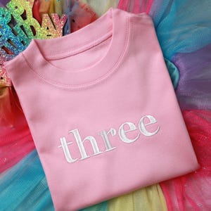 May include: Pink t-shirt with the word "three" embroidered in white on the front. The shirt is lying on a rainbow tulle fabric.