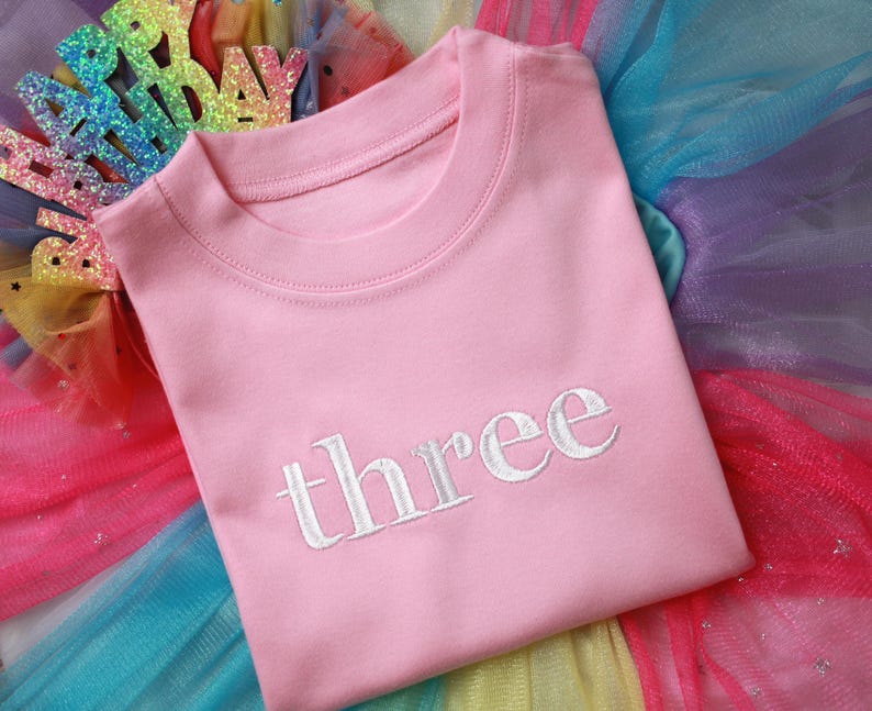 May include: Pink t-shirt with the word "three" embroidered in white on the front. The shirt is lying on a rainbow tulle fabric.