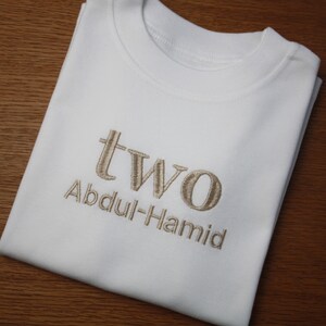 May include: White t-shirt with the word "two" and the name "Abdul-Hamid" embroidered in brown thread.