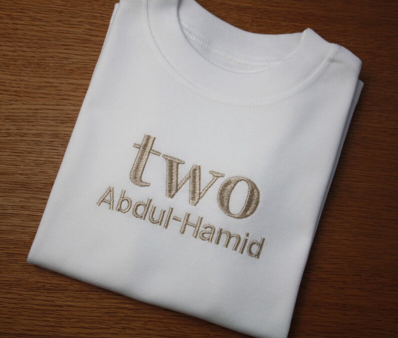 May include: White t-shirt with the word "two" and the name "Abdul-Hamid" embroidered in brown thread.
