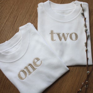 May include: Two white t-shirts with the words "one" and "two" embroidered in beige on the front.