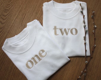 May include: Two white t-shirts with the words "one" and "two" embroidered in beige on the front.