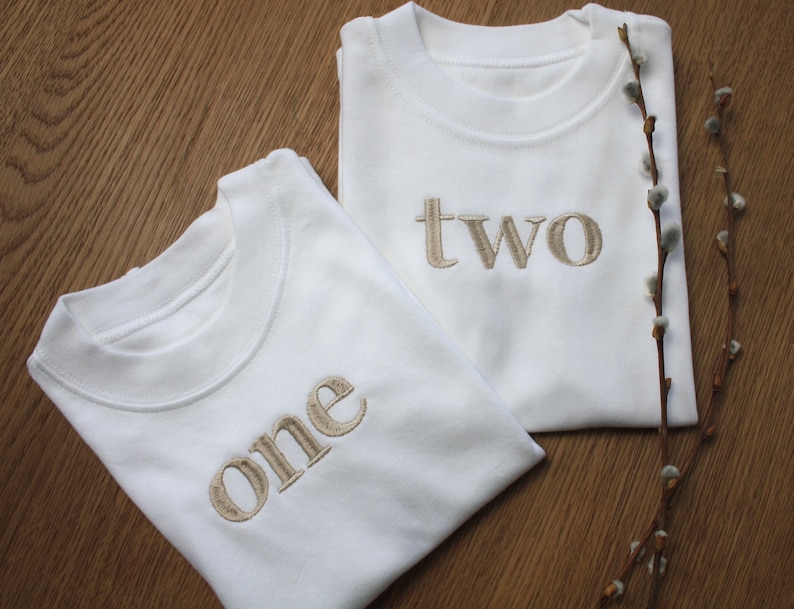May include: Two white t-shirts with the words "one" and "two" embroidered in beige on the front.