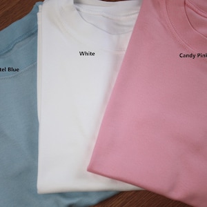 May include: Three plain t-shirts in pastel blue, white, and candy pink. The shirts are folded and stacked on top of each other. The colours of the shirts are labelled with text.