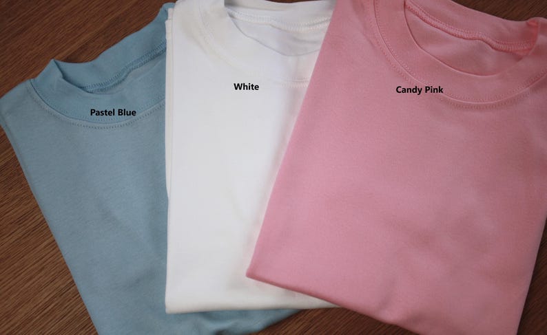 May include: Three plain t-shirts in pastel blue, white, and candy pink. The shirts are folded and stacked on top of each other. The colours of the shirts are labelled with text.