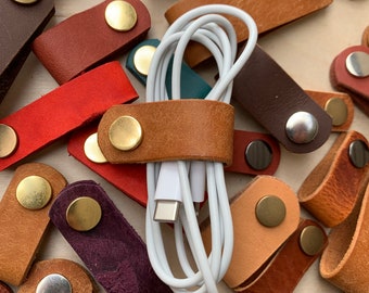 May include: A collection of leather cord organizers in various colors and styles, with a white cord running through one of the organizers. The organizers have brass and silver metal accents.